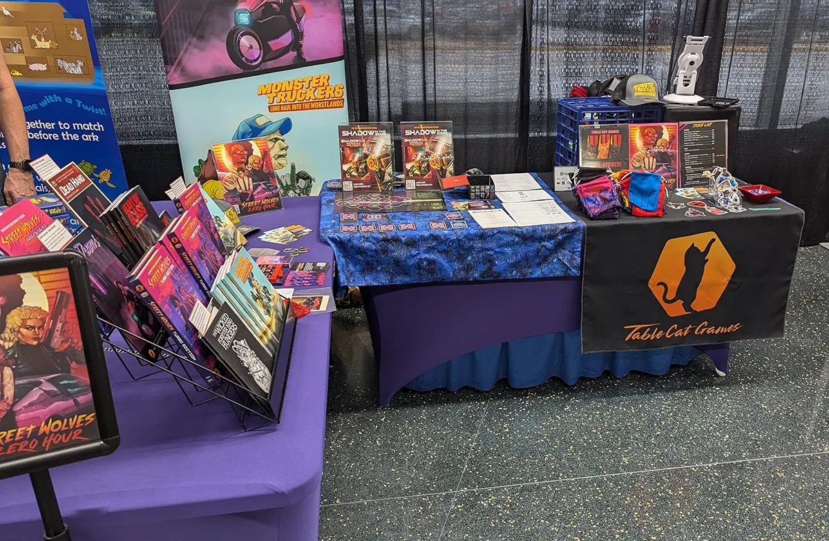 Image of a booth at a convention that is an L shape. It has the Table Cat Games banner on it along with all the games and stuff displayed in a nice fashion.